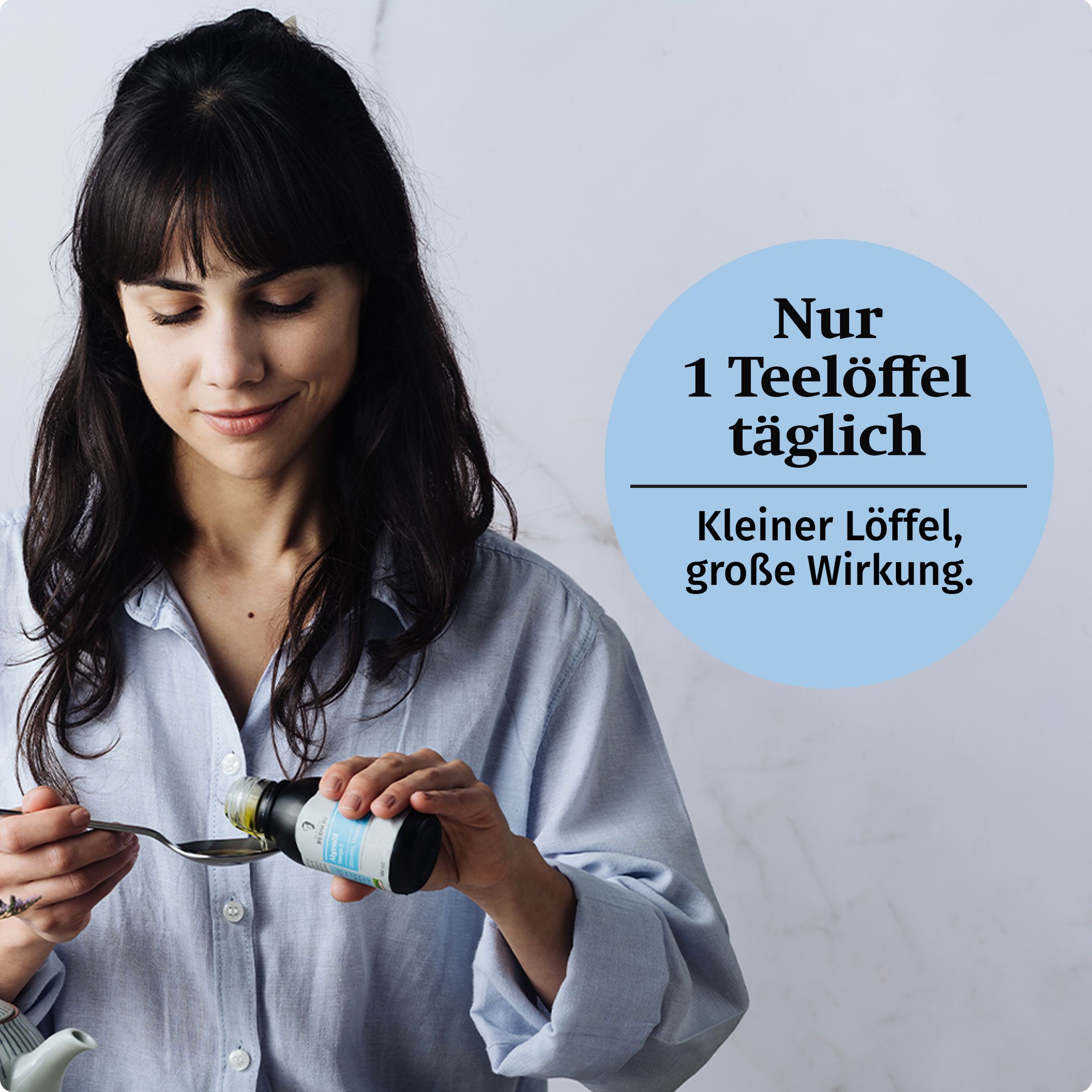Frau mit schwarzen Haaren hält Dr.Budwig Algenöl in der Hand und kippt es auf einen Löffel. Rechts darüber ist ein blaufarbiger Kreis mit dem Schriftzug: Nur 1 Teelöffel täglich. Kleiner Löffel, große Wirkung.